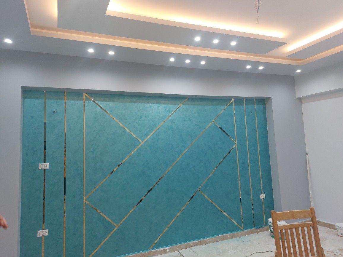 An interior being prepared: a turquoise textured wall panel divided by masking-tape guidelines, beneath a finished false ceiling with cove lighting and downlights.