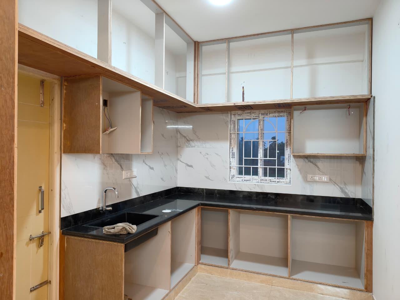A kitchen mid-installation: timber base and wall units fitted under a dark countertop, with the window frame still masked and the units not yet finished.