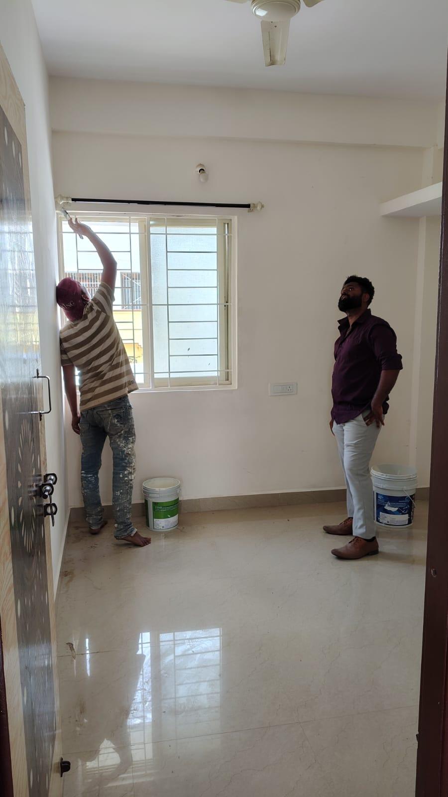 Two Payyonpaints painters working in a room: one reaches up with a roller to cut in along a window reveal while the other watches on, with open paint buckets on the floor.