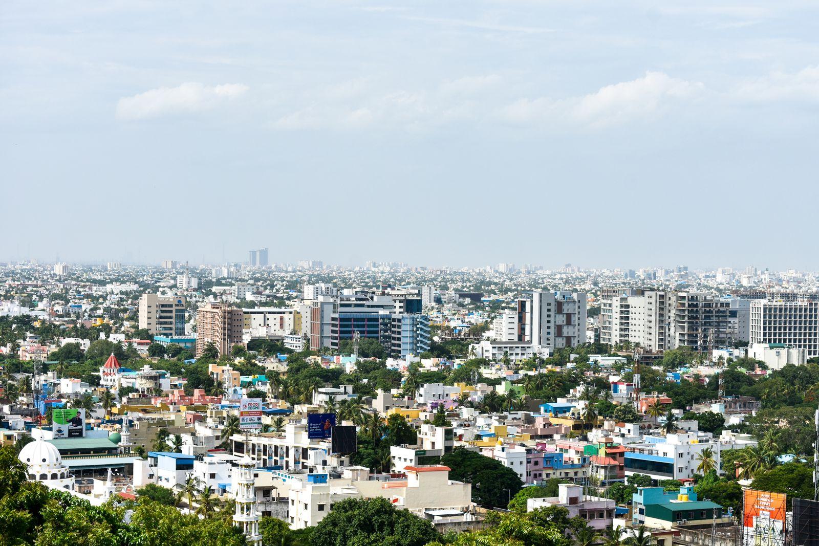 Aerial view across Chennai: dense low-rise rooftops, palms and mid-rise blocks stretching to the horizon under a bright sky